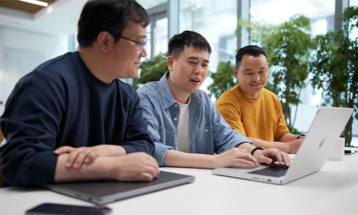 Ken escribe en su MacBook, otras personas del equipo están sentadas junto a él, hay ventanas con iluminación y plantas al fondo.