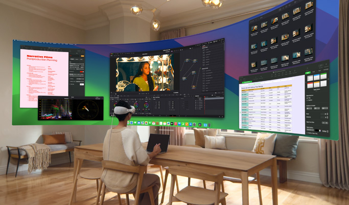 A woman sitting at a desk wearing Apple Vision Pro. In front of her, in 3D space, are mulitple screens each with a different app in use.