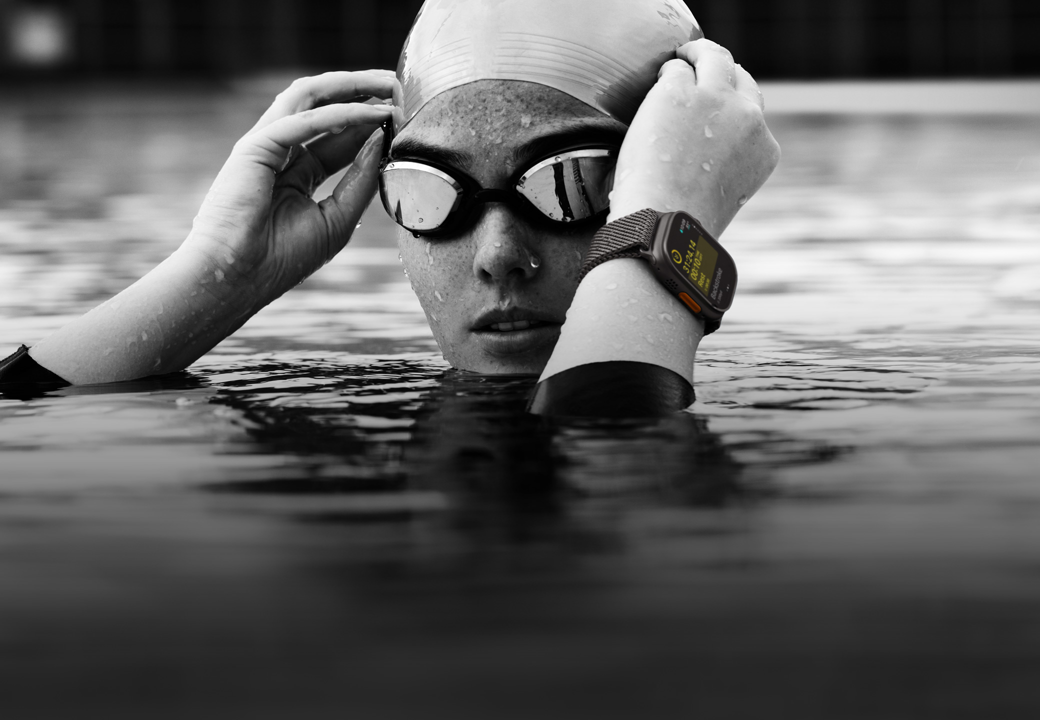 En svømmer med hovedet og hænderne over vandet i en swimmingpool. Svømmeren har et Apple Watch Ultra 2 på og retter på sine svømmebriller.