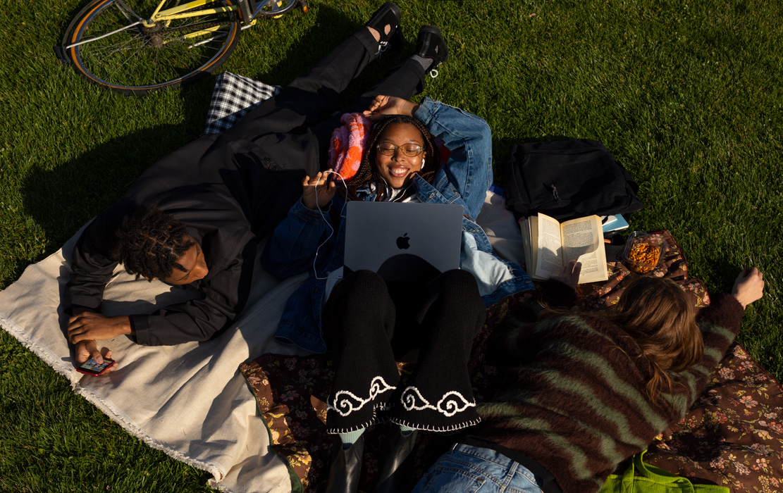Tres estudiantes universitarios acostados sobre una manta en el parque. Un estudiante tiene un iPhone. Una estudiante tiene una MacBook Air y audífonos Apple. Una estudiante tiene un libro de bolsillo.