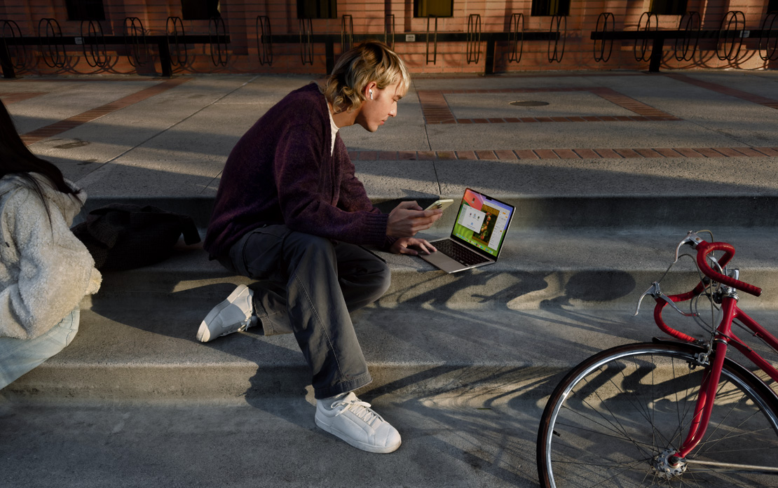 Un estudiante universitario sentado en unas escaleras usa un iPhone y una MacBook. A su lado, una bicicleta.