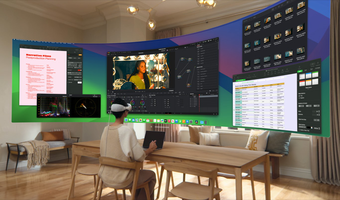 A woman sitting at a desk wearing Apple Vision Pro. In front of her, in 3D space, are mulitple screens each with a different app in use.