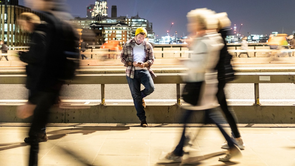 Ein Mann steht im Großstadttrubel auf einer Brücke und schaut auf sein Handy. Viele Menschen treibt die Angst um, in ihrer Freizeit etwas zu verpassen. Wir haben Tipps gegen FOMO und Freizeitstress, damit ihr euch in eurer freien Zeit wirklich erholt. | Bild: picture alliance / Westend61 | William Perugini