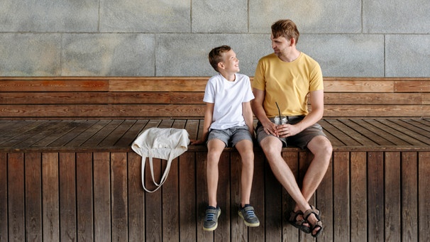 Vater und Sohn unterhalten sich, beide sitzen auf einer Holzbank. Was bedeutet Gewaltfreie Kommunikation mit Kindern? Der Psychologe Marshall B. Rosenberg hat sie entwickelt. Wir erklären, warum Gewaltfreie Kommunikation mit Kindern wichtig ist, wie ihr sie in vier Schritten erlernen könnt und was es mit der Giraffensprache auf sich hat.  | Bild: picture alliance/Westend61/Ekaterina Yakunina