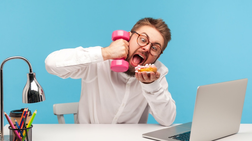 Zu viel Multitasking ist nicht gesund. Es verursacht Stress und kann auf Dauer zu einem Burnout oder zu einer Depression führen. Im Bild: Ein Mann sitzt am Schreibtisch vor dem Computer, hält in einer Hand eine Hantel in der andern ein Stück Kuchen. | Bild: picture alliance / Zoonar | Khosrow Rajab Kordi