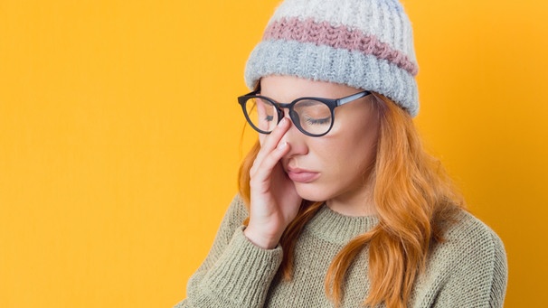 Eine müde wirkende junge Frau mit Brille und Mütze hält sich mit einer Hand die Augen zu. Krisen aushalten und überwinden - und dabei leistungsfähig bleiben. Unzählige Ratgeber behaupten, das ließe sich trainieren. Wie sinnvoll ist das? Setzen wir uns damit nicht erst recht unter Druck? | Bild: colourbox.com