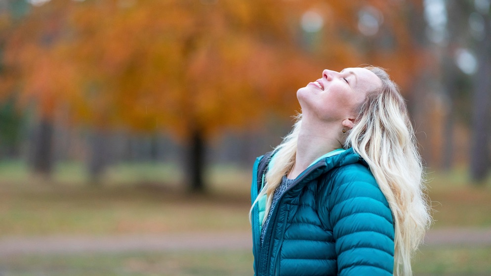 Eine Frau steht im Park und lächelt mit geschlossenen Augen. Selbstliebe bedeutet, mit sich selbst befreundet zu sein, zu seinen Bedürfnissen und Wünschen zu stehen und die eigenen Schwächen zu akzeptieren. Wie du lernst, dich selbst zu lieben. | Bild: picture alliance / Westend61 | William Perugini