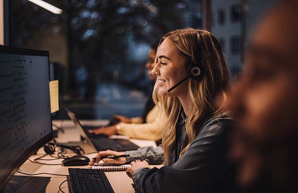 Smiling blond female customer service representative wearing headset using computer at desk in call center