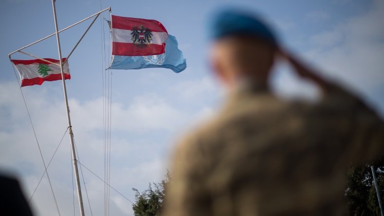 Das Bild zeigt im Hintergrund die österreichische, die libanesische und die UN Flagge an einer Fahnenstange. Die Flaggen sind im Fokus. Im Vordergrund ist der Umriss eines Soldaten zu sehen, der salutiert.