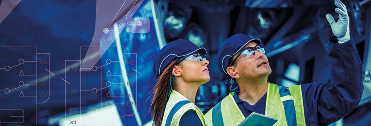 Two people in bump caps, safety glasses and ear protection look up at an engine