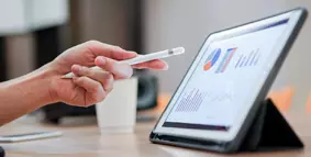 a close up of a hand holding a pen pointing at a tablet with graphs on the screen