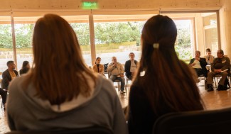 Two women (back facing the viewer) in foreground look onto a meeting of people seated around a large table.