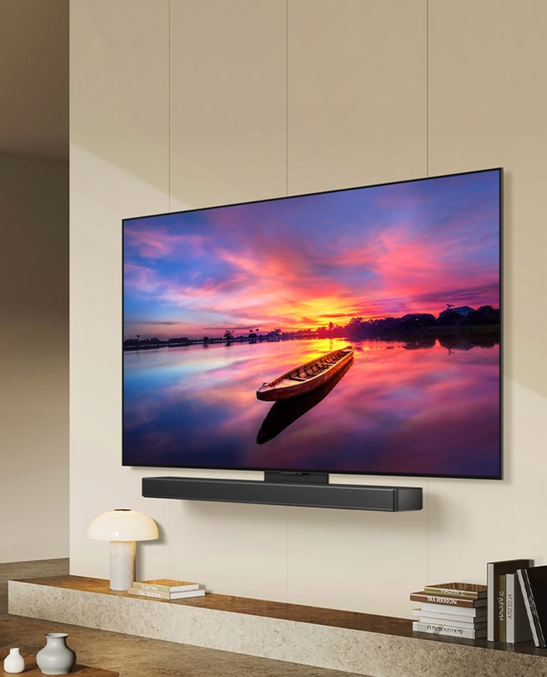 Uma imagem da LG OLED C4, voltada 45 graus para a esquerda, exibindo um belo pôr-do-sol com um barco em um lago, com a TV conectada a uma LG soundbar por meio do suporte Synergy em uma sala de estar moderna.