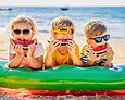 Drei Kinder im Grundschulalter liegen bäuchlings auf einer Luftmatratze am Strand. Ihre Gesichter sind durch Sonnenbrillen und Melonenschnitze vor dem Mund teilweise verdeckt.