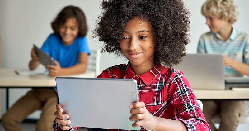 Eine Schüler sitzt lächelnd vor einem Tablet in einem Klassenzimmer.