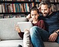 Ein Vater sitzt neben seiner Tochter auf einem Sofa. Die Tochter hält ein Tablet in der Hand und gestikuliert.