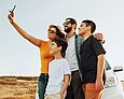 Eine Frau hält ein Handy in die Höhe, um ein Selfie mit ihrem Mann und ihren zwei Söhnen zu machen. Sie stehen vor einem weißen Auto in einer trockenen Steppenlandschaft.