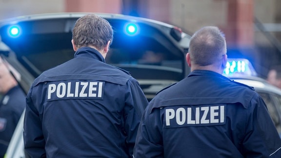 Zwei Polizisten mit dem Schriftzug Polizei auf dem Rücken der Dienstjacke hinter einem Streifenwagen mit Blaulicht