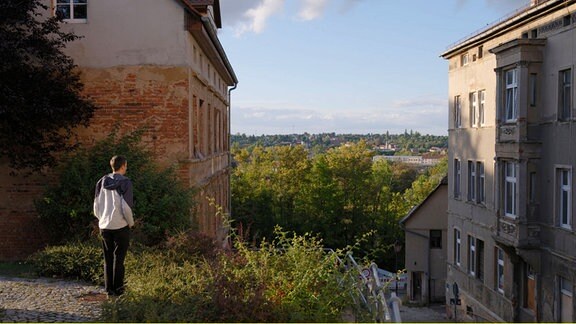 Ein Mann steht auf einem Plateau und blickt auf Häuser und Bäume, im Hintergrund blauer Himmel. 