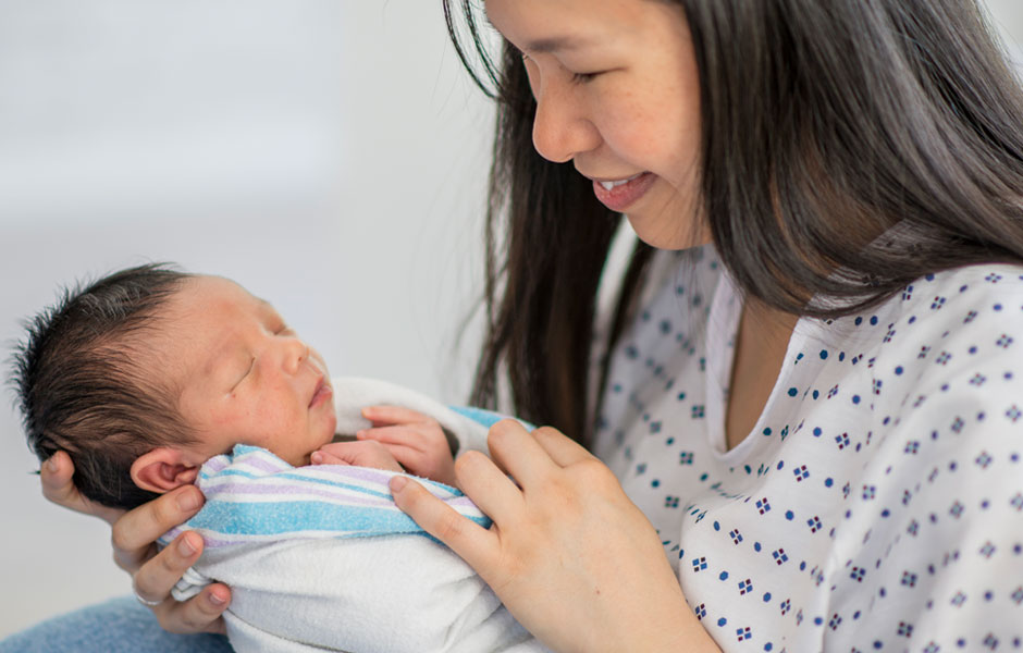 Mother in hospital gown smiles at her newborn who is swaddled in a hospital blanket.  