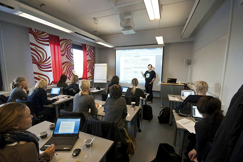 Et klasserom med en foreleser og studenter med bærbare datamaskiner. Rommet har hvite vegger, røde gardiner, og en tavle på venstre side.