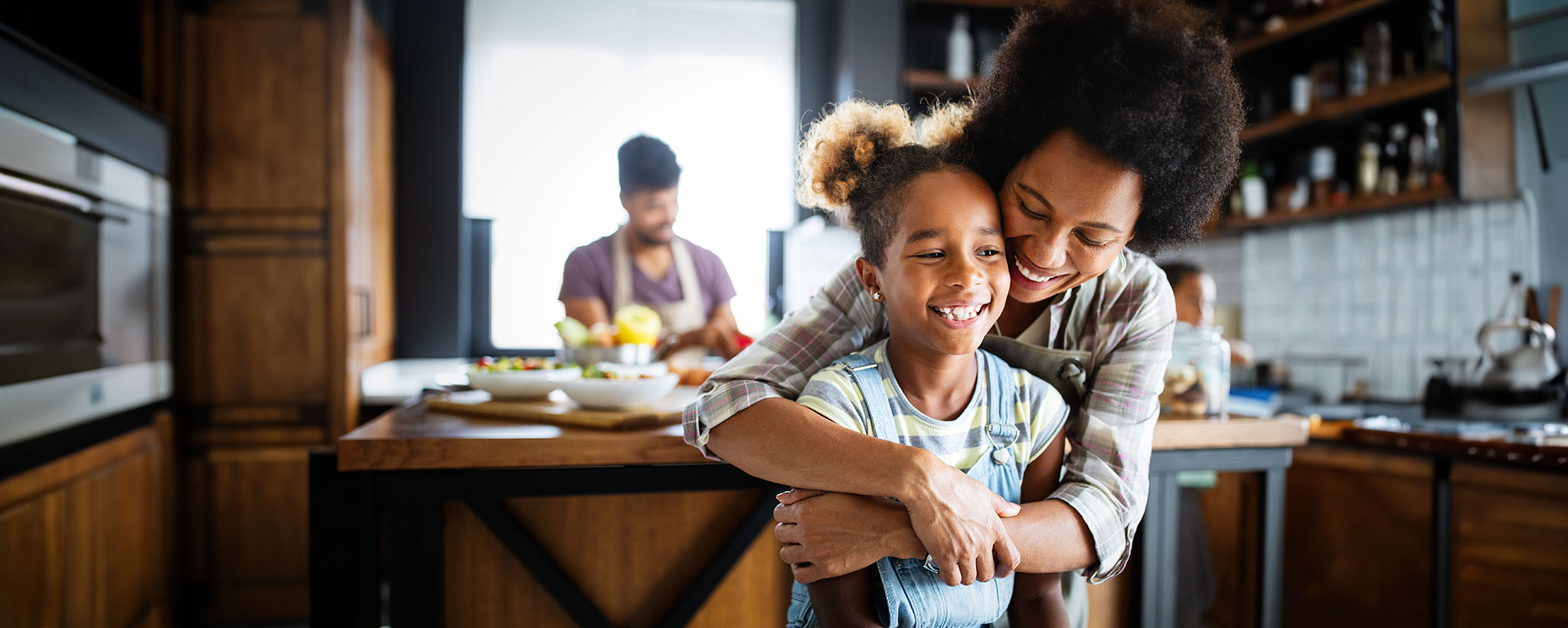 Mother hugs daughter in a kitchen as dad and sibling cook in the background. 