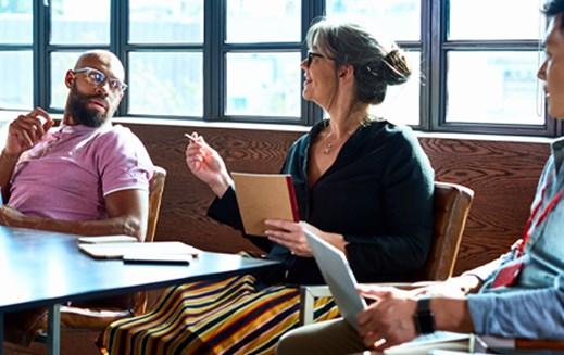 Colleagues engaged in conversation around a table. The woman speaking gestures with her pen.