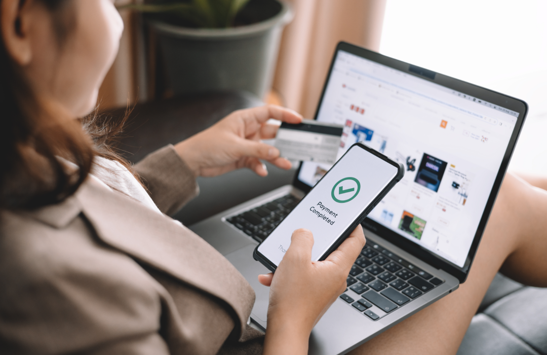 Woman using a laptop to shop online while holding a credit card and confirming payment on her smartphone