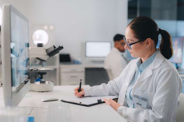 Woman in lab coat writing in notebook on desk