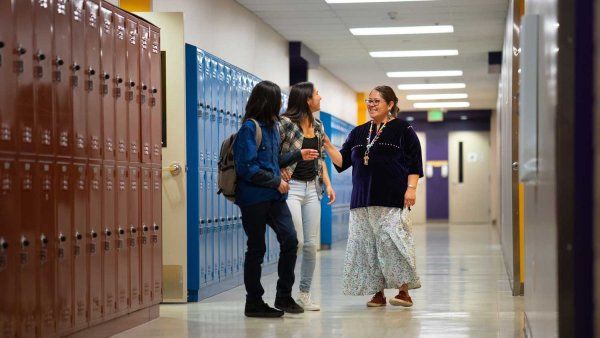 A teacher laughs with two female high school students at a school hallway.