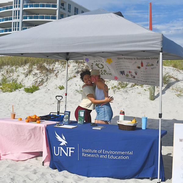 Two students standing under a tent on the beach, hugging and smiling 