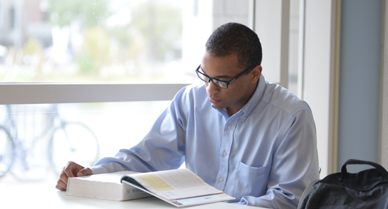 Male student at a desk reading a large text book