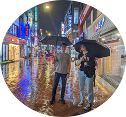 two UNF students with umbrellas smiling on a rainy night street with lights in Seoul