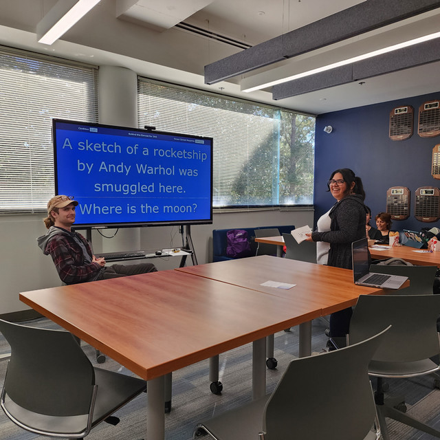 Two students stand in front of a tv with a Trivia question on it.