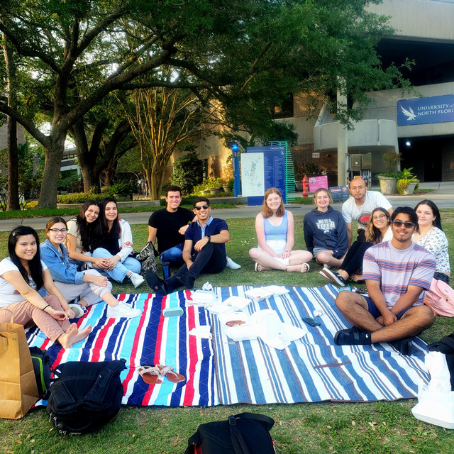 A group of students sit around a patterned blanket on a large patch of grass.