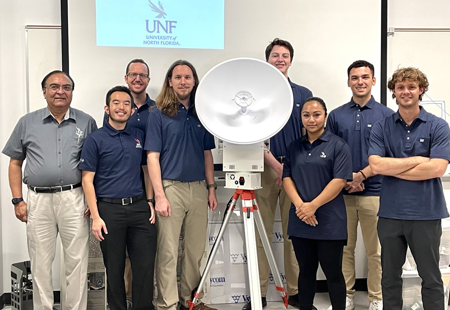 UNF student team and instructor Dr. Nirmal Patel that is participating in two NASA research projects posing in a group photo next to a satellite