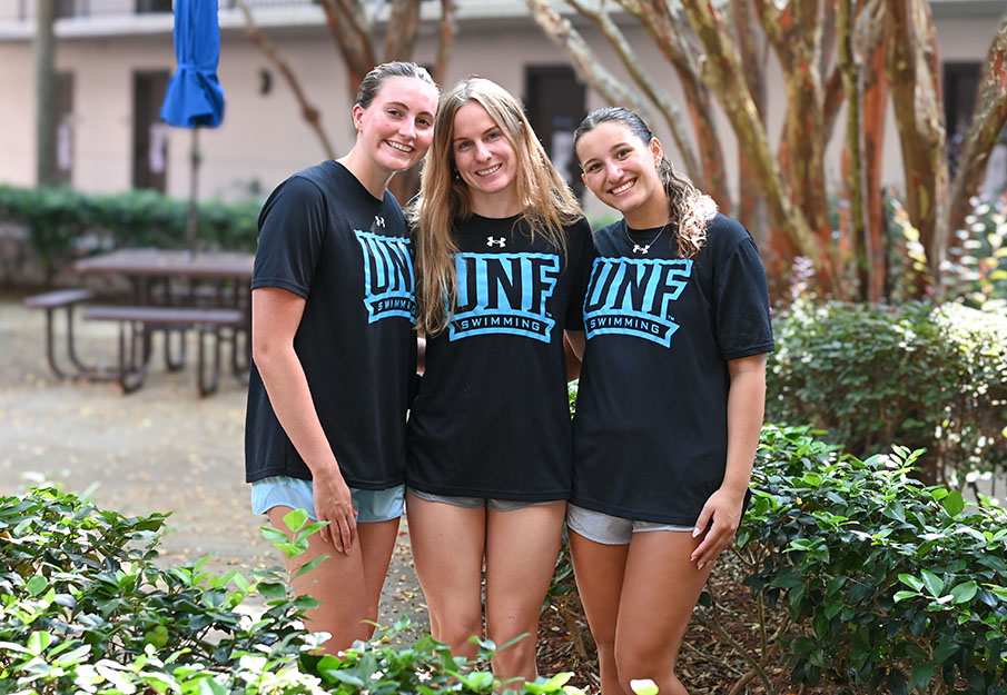 Three UNF swim team girls posed together in front of one of the residence halls