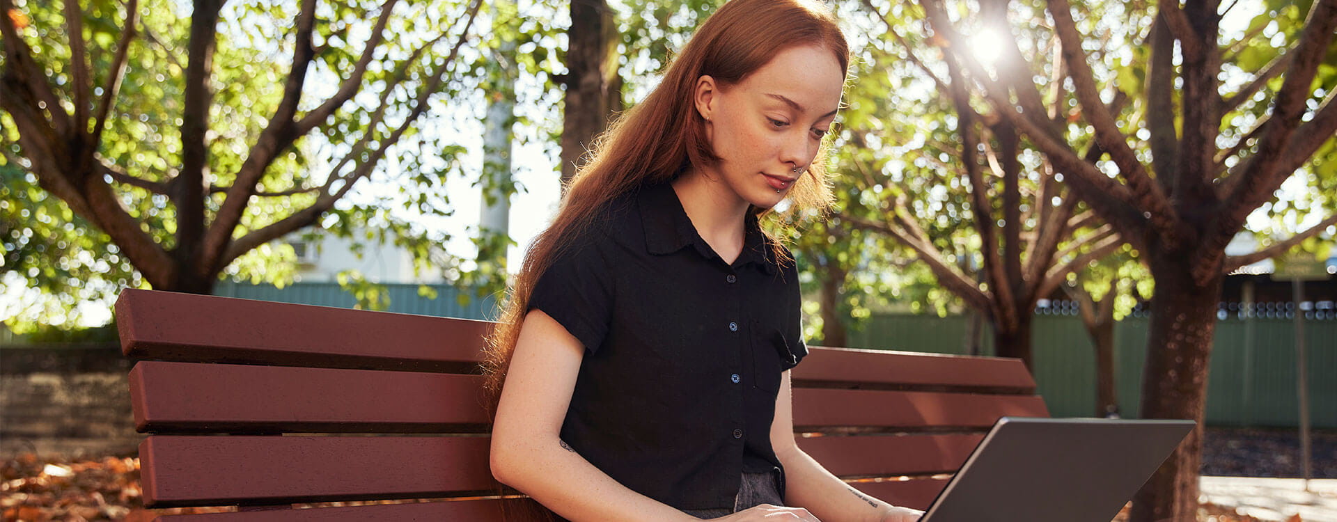 A person with long hair sits on an outdoor bench using a laptop. Trees and sunlight are in the background.