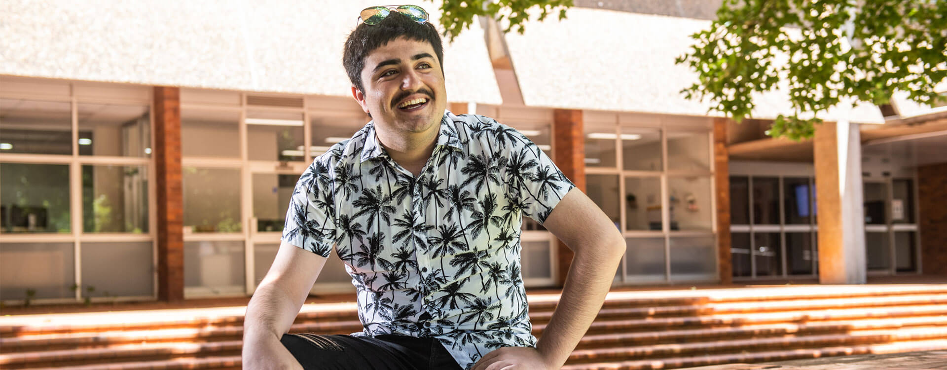 A man with a mustache and wearing a palm tree-patterned shirt smiles while sitting on steps in front of a building with large windows.