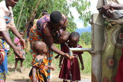 A woman helps children drink water from a tap outside.