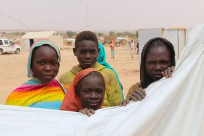 Three women and a young man look at the camera. Some of the women are clutching a plastic sheet. A UN vehicle and tents can be seen in the background.