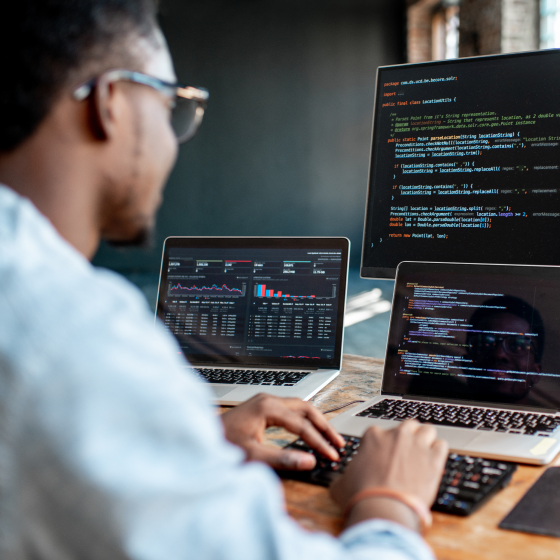 A black man with glasses and wearing a blue shirt sitting at a desk with a keyboard and multiple screens showing code and reports