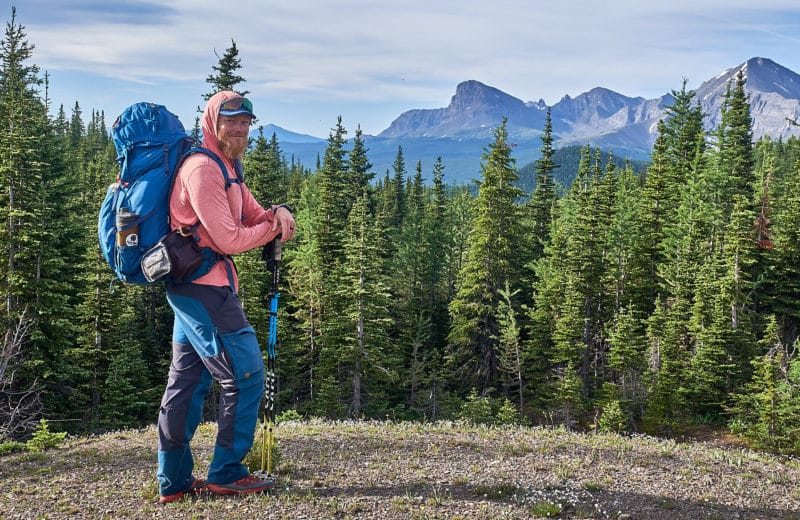 Man with blue backpack and hiking poles standing in front of a picturesque alpine landscape