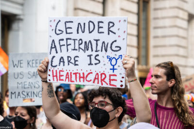 A shirtless demonstrator (wearing glasses and a black surgical mask) holds up a sign that reads" GENDER AFFIRMING CARE IS HEALTHCARE".