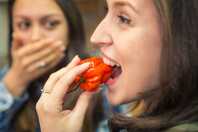 A woman eats a red chili pepper, while a shocked friend watches