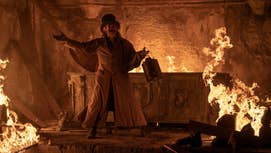 Willem Dafeo in 2024's Nosferatu, he's dressed in old attire, holding some kind of metal container stood in what looks like a mausoleum, which is partially on fire.