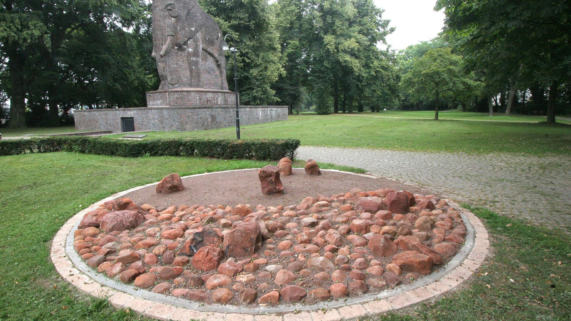 Das Bremer Herero-Mahnmal, das an die Opfer der deutschen Kolonialherrschaft im heutigen Namibia erinnern soll. Man sieht einen Steinkreis mit einzelnen herausgehobenen Brocken, dahinter das Antkolonialismus-Mahnmal, dass einen Steinelefanten zeigt.