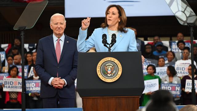 Archivbild: Kamala Harris steht bei einer Wahlkampfveranstaltung am Rednerpult und spricht zum Publikum, links hinter ihr steht Joe Biden. Im Hintergrund sind Unterstützer der Demokraten mit Wahlkampfplakaten in den Händen zu sehen.