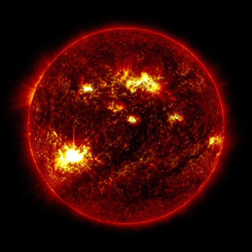 The Sun, shown in red, appears against a black background. Several bright yellow active regions appear across the Sun. A bright flash of yellow and white light can be seen in the lower left part of the Sun.