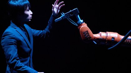 Theatrically lit person with their arm up, moving to touch a robot arm